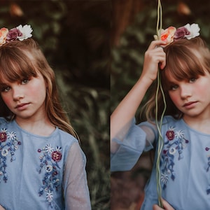 Può includere: Una ragazza giovane con lunghi capelli castani indossa un vestito azzurro con ricami floreali e una corona di fiori. Tiene un tralcio verde in mano.
