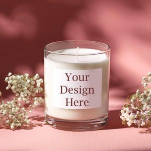 May include: A white candle in a clear glass jar with a label that says "Your Design Here" on a pink surface with white flowers.