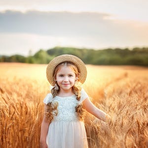 May include: A young girl wearing a white dress and a straw hat smiles while standing in a field of golden wheat.