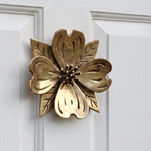 May include: A polished brass door knocker shaped like a five-petaled flower with detailed veining on each petal and a central cluster of small rounded stamens. The metallic floral ornament is mounted centrally on a white paneled door.