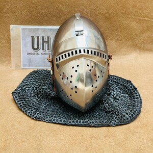 Op de afbeelding: Een zilveren helm met een gezichtsmasker en een kettingbaard. De helm heeft een scharnierend vizier en een leren band. De helm staat op een bruin oppervlak.