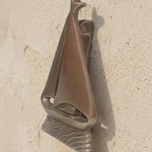 May include: A bronze-colored sailboat door knocker. The door knocker is mounted on a textured, light-colored wall. The sailboat design features a detailed hull and sail, with a wave pattern at the base.