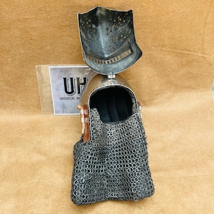Op de afbeelding: Een middeleeuwse helm met een kettingbaard, de helm is gemaakt van metaal en heeft een scharnierend vizier dat omhoog is geklapt. De helm staat op een bruin oppervlak.