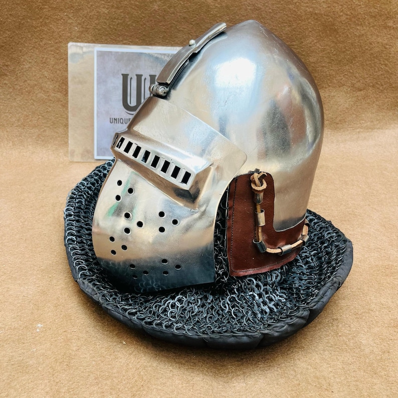 Op de afbeelding: Een zilveren helm met een vizier en een kettingbaard. De helm heeft een bruin leren band en een metalen gesp. De helm staat op een zwarte achtergrond.