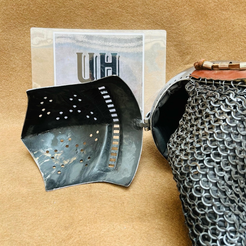 Op de afbeelding: Een close-up van een middeleeuwse helm met een kettingbaard. De helm is gemaakt van metaal en heeft een vizier dat kan worden opgetild en neergelaten. De kettingbaard is gemaakt van kleine metalen ringen die aan elkaar zijn verbonden.