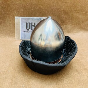 Op de afbeelding: Een zilveren helm met een afgeronde bovenkant en een zwarte gepolsterde basis. De helm staat op een bruin oppervlak. De tekst "UNIQUE CAL HAT" is zichtbaar op een wit label achter de helm.