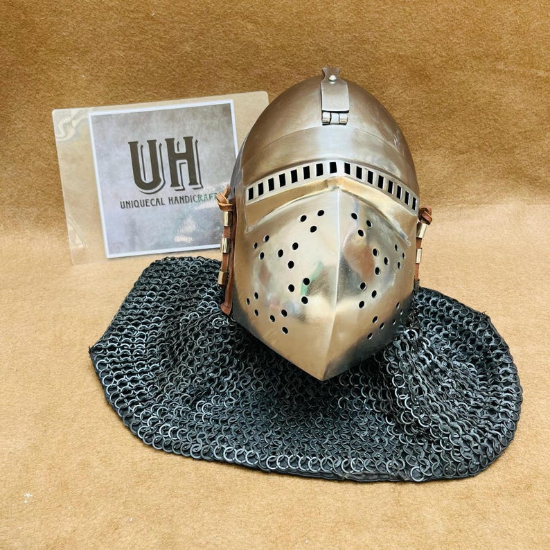 Op de afbeelding: Een zilveren helm met een vizier en een kettingbaard. De helm heeft een reeks kleine gaten in het vizier en een decoratieve band rond de bovenkant. De helm staat op een bruin oppervlak.  "UH UNIQUE CAL HANDICRAFT" is afgedrukt op een wit label.