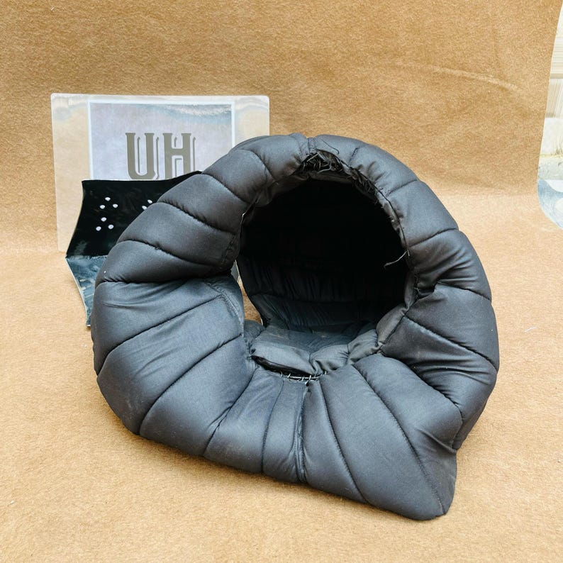 Op de afbeelding: Een zwarte gewatteerde helm met een ronde opening aan de bovenkant. De helm is gemaakt van een zacht, gevoerd materiaal.