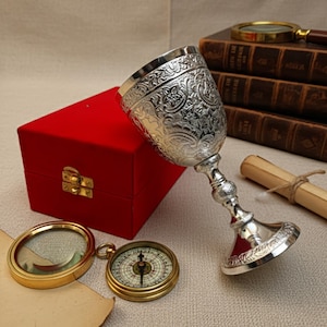 May include: A silver goblet with ornate floral patterns, resting beside a red velvet box, a compass, and a magnifying glass. A rolled parchment and antique books are in the background. The goblet has a detailed stem and base.