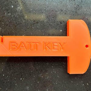 Könnte beinhalten: Hellorangefarbenes "BAIT KEY" Werkzeug. Das Werkzeug hat die Form eines T mit einem rechteckigen Griff und einer abgerundeten Oberseite. Ein kleines Loch ist in die abgerundete Oberseite gebohrt. Der Text "BAIT KEY" ist auf dem Griff geprägt.