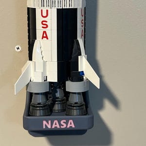 May include: A model rocket constructed from white and black building blocks, featuring the red text USA vertically on the side. The base is held in a dark gray mount with the word NASA printed in pink letters, showcasing detailed engine nozzles.