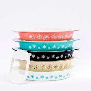 May include: A stack of vintage Pyrex oval baking dishes in various colors: coral, turquoise, black, cream, and white. Each dish features a decorative floral or snowflake pattern in white or turquoise. A white plastic lid is partially visible.