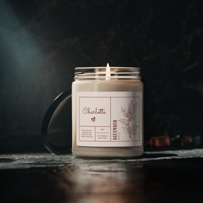 K&ouml;nnte beinhalten: Eine wei&szlig;e Kerze in einem Glasgef&auml;&szlig; mit einem braunen Etikett. Das Etikett tr&auml;gt den Text "Charlotte" mit einem Herzsymbol und "December Holly" mit einer Zeichnung von Stechpalmenbl&auml;ttern. Die Kerze ist angez&uuml;ndet und hat einen warmen Schein.