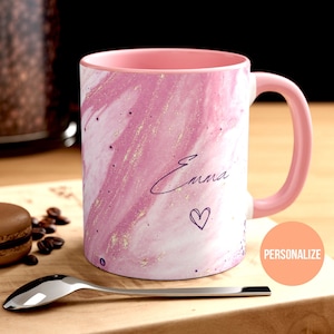 May include: Pink and white marble mug with gold glitter accents. The mug has a pink rim and handle. The name "Emma" is written in purple script on the mug with a small heart below it.