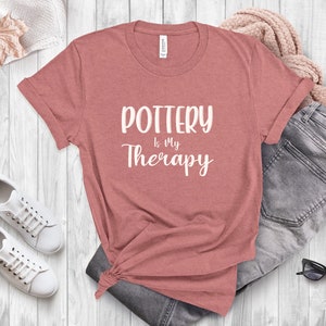 May include: A heather pink t-shirt with the text "POTTERY is My Therapy" in white lettering.