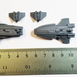 K&ouml;nnte beinhalten: Ein Satz von f&uuml;nf grauen Plastik-Raumschiffmodellen, darunter ein gr&ouml;&szlig;eres Schiff mit zwei Triebwerken und ein kleineres Schiff mit zwei Fl&uuml;geln. Die Modelle sind auf einer wei&szlig;en Oberfl&auml;che mit einem Lineal zur Skalierung angeordnet.