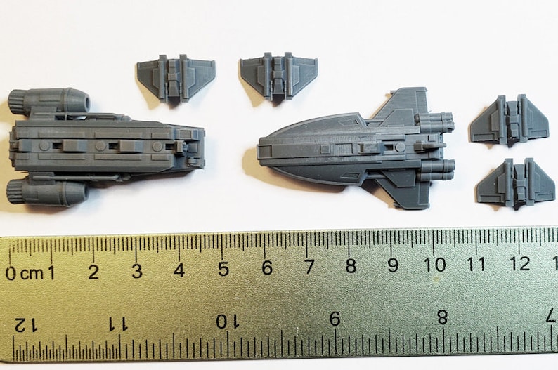K&ouml;nnte beinhalten: Ein Satz von f&uuml;nf grauen Plastik-Raumschiffmodellen, darunter ein gr&ouml;&szlig;eres Schiff mit zwei Triebwerken und ein kleineres Schiff mit zwei Fl&uuml;geln. Die Modelle sind auf einer wei&szlig;en Oberfl&auml;che mit einem Lineal zur Skalierung angeordnet.
