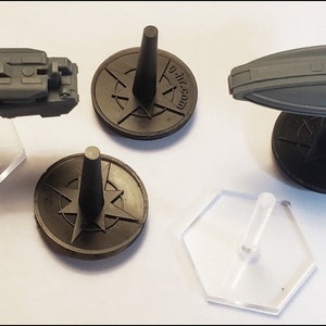 K&ouml;nnte beinhalten: Drei Miniatur-Raumschiffe, zwei auf schwarzen Sockeln mit einem Kompass-Design und eines auf einem durchsichtigen Plastiksockel. Die Raumschiffe sind grau und haben detaillierte Merkmale.