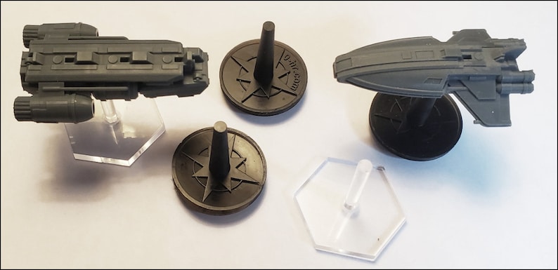 K&ouml;nnte beinhalten: Drei Miniatur-Raumschiffe, zwei auf schwarzen Sockeln mit einem Kompass-Design und eines auf einem durchsichtigen Plastiksockel. Die Raumschiffe sind grau und haben detaillierte Merkmale.
