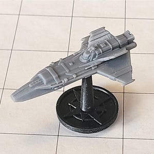 May include: A grey plastic miniature spaceship model featuring a pointed nose, angular wings, and a detailed fuselage. The spacecraft is mounted on a black circular stand with a star-shaped base, designed for tabletop gaming and display purposes.