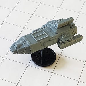 May include: A gray plastic spaceship model with a detailed design, featuring a cockpit, engines, and a weapon system. The model is mounted on a black base.