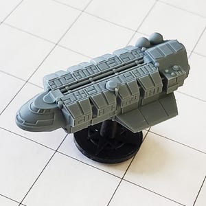 May include: A gray, detailed, 3D printed model of a spaceship with a rounded nose and a long, rectangular body. The model is mounted on a black base.