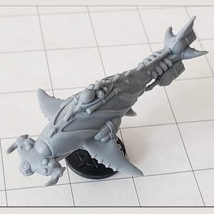 Op de afbeelding: Een grijs, gedetailleerd miniatuur ruimteschipmodel met een ontwerp dat lijkt op een hamerhaai. Het model heeft vinnen, een getextureerd oppervlak en diverse mechanische details. Gemonteerd op een zwarte, ronde basis.