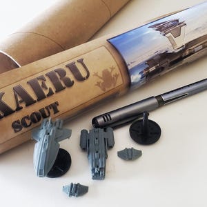 K&ouml;nnte beinhalten: Eine braune Pappr&ouml;hre mit dem aufgedruckten Text "KAERU SCOUT". Die R&ouml;hre enth&auml;lt einen Satz von f&uuml;nf grauen, kleinen Raumschiffen aus Plastik.