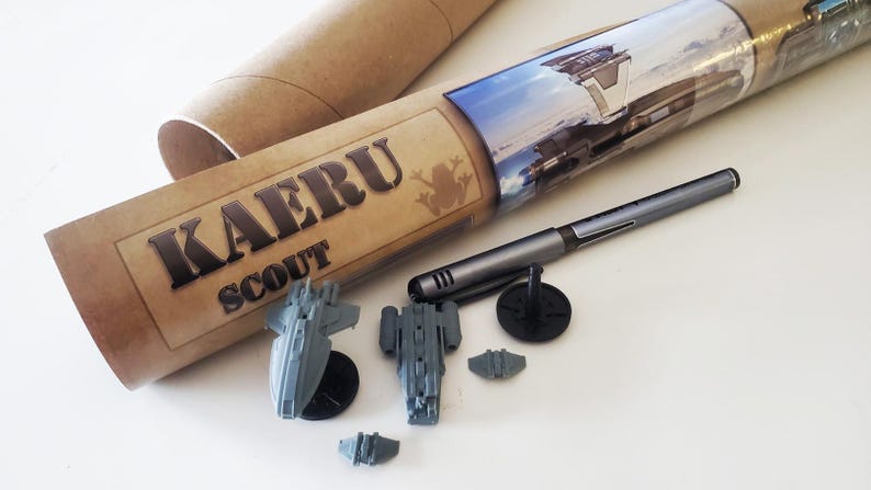 K&ouml;nnte beinhalten: Eine braune Pappr&ouml;hre mit dem aufgedruckten Text "KAERU SCOUT". Die R&ouml;hre enth&auml;lt einen Satz von f&uuml;nf grauen, kleinen Raumschiffen aus Plastik.