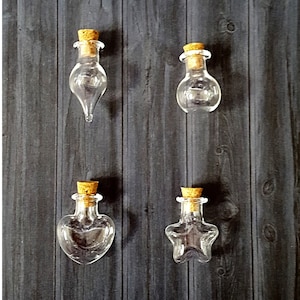 May include: Four small glass bottles with cork stoppers. The bottles are shaped like a teardrop, a sphere, a heart, and a star. The bottles are clear and empty.