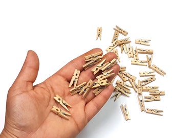 May include: A collection of small, natural wood clothespins with silver-colored metal springs. Some clothespins are held in an open palm, while others are scattered on a white surface. The clothespins are approximately 1 inch in length, ideal for crafts or displaying photos.