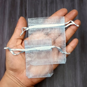 May include: Two light blue sheer organza gift bags with silver ribbon ties. The bags are nested inside each other.