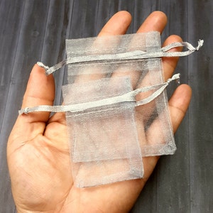 May include: Two small, sheer silver organza drawstring bags. The bags are rectangular with a delicate, shimmering texture. Each bag has a matching silver satin ribbon drawstring closure. The bags are shown against a neutral background.