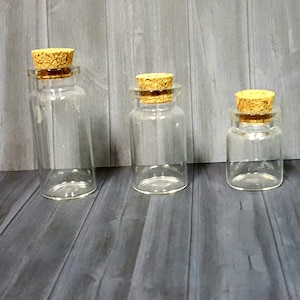 May include: Three clear glass bottles with cork stoppers. The bottles are different sizes and are arranged in a row from largest to smallest.