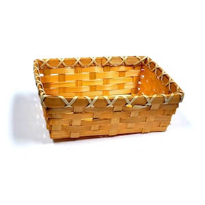 May include: A rectangular woven basket made of light brown bamboo. The basket features a crisscross pattern and is trimmed with a lighter-colored material along the top edges. The basket is empty and sits on a white background.