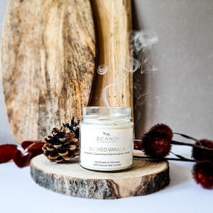 Könnte beinhalten: Eine durchsichtige Glaskerze in einem Glas mit einem weißen Etikett, auf dem "SCANDI SMOKED VANILLA Bergamot | Caramelized Marshmallows | Vanilla Handmade in Germany 100% Natural Wax Candles" steht. Die Kerze ist angezündet und Rauch steigt aus der Oberseite auf.