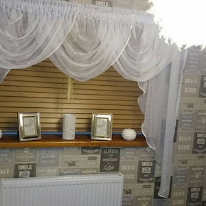 Puede incluir: Cortinas blancas y transparentes con un borde festoneado cuelgan de una ventana con persianas de madera. Las cortinas se juntan en el centro para crear un efecto de festón. Hay tres objetos decorativos en el alféizar de la ventana, incluyendo dos imágenes enmarcadas y una vela.