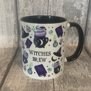 May include: White ceramic mug with a black handle and black rim. The mug has a purple, green, and black design with a cauldron, stars, and the text "Witches Brew".