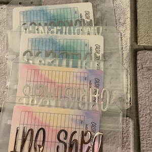 May include: Clear plastic cash envelope system with silver lettering and colorful watercolor-style inserts. The envelopes are labeled with various spending categories. The background is a textured, neutral-toned surface.