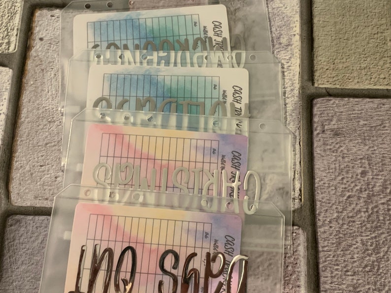 May include: Clear plastic cash envelope system with silver lettering and colorful watercolor-style inserts. The envelopes are labeled with various spending categories. The background is a textured, neutral-toned surface.