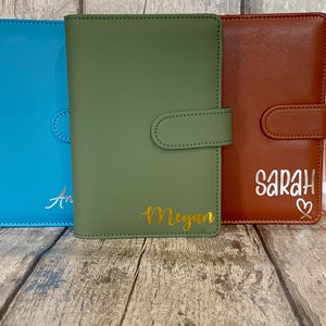 Op de afbeelding: Vijf gepersonaliseerde lederen notitieboekjes in verschillende kleuren, waaronder groen, blauw, oranje, rood en bruin. De notitieboekjes hebben een klep sluiting en zijn gepersonaliseerd met de namen "Megan", "Sarah" en "Ann".