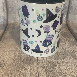 May include: White ceramic mug with a black rim and handle. The mug features a repeating pattern of witch hats, cauldrons, moons, stars, and other witchy imagery. The text "Witchy Vibes" is printed on the mug.