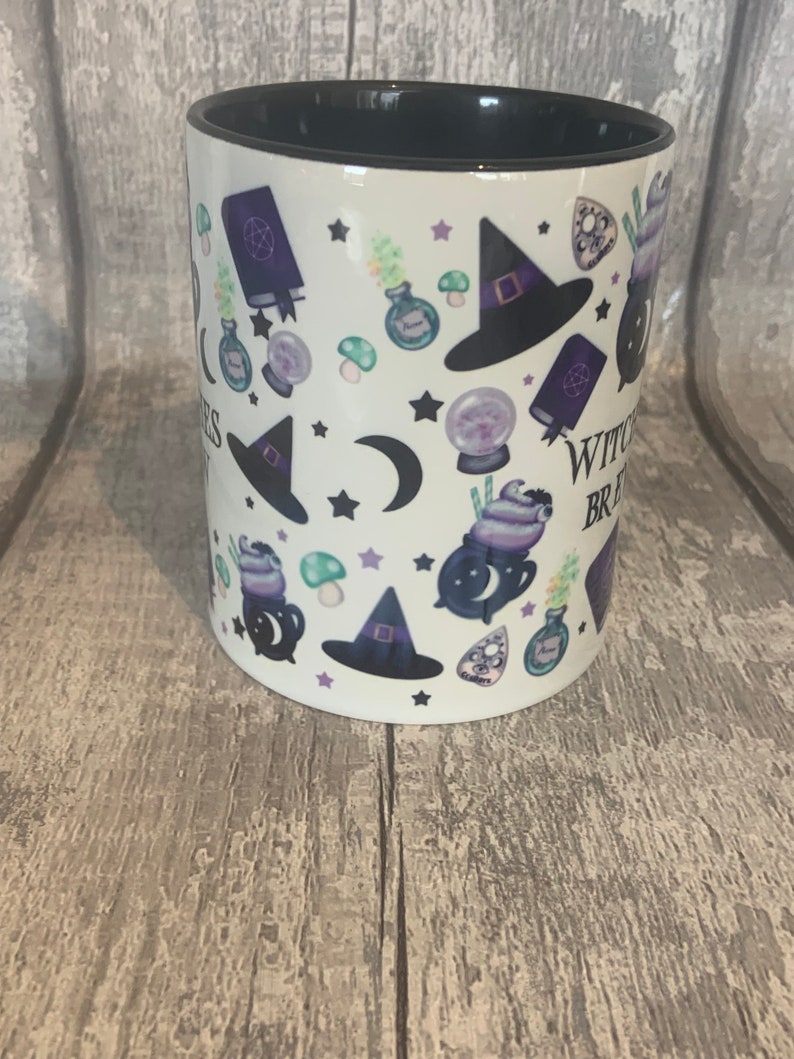 May include: White ceramic mug with a black rim and handle. The mug features a repeating pattern of witch hats, cauldrons, moons, stars, and other witchy imagery. The text "Witchy Vibes" is printed on the mug.