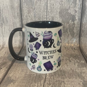May include: White ceramic mug with a black handle and a black rim. The mug has a design of a cauldron, a witch's hat, stars, and other witchy elements. The text "Witches Brew" is printed on the mug.
