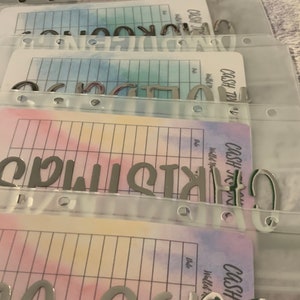 May include: Clear plastic cash envelope system with labels. The labels read "Christmas", "Groceries", and "Clothing". Each envelope has a colorful watercolor design and a grid for tracking expenses.
