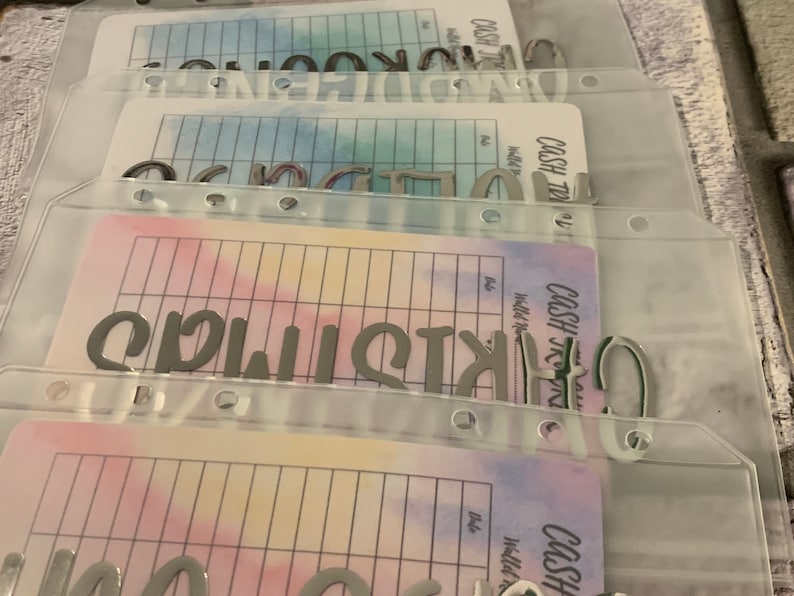 May include: Clear plastic cash envelope system with labels. The labels read "Christmas", "Groceries", and "Clothing". Each envelope has a colorful watercolor design and a grid for tracking expenses.
