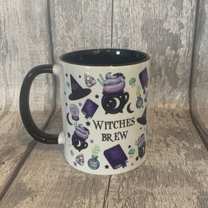 May include: White ceramic mug with a black handle. The mug has a black rim and features a white background with a black and purple design of a cauldron, stars, a witch's hat, and the text "Witches Brew".