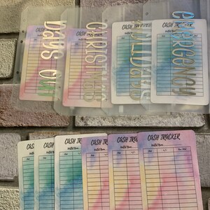 May include: A collection of cash tracker inserts and dividers. The inserts feature watercolor backgrounds in pastel hues. The dividers have metallic lettering spelling out "Days Off", "Christmas", "Wedding", and "Emergency".