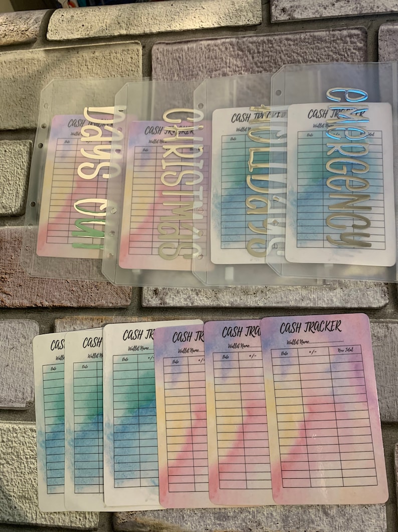 May include: A collection of cash tracker inserts and dividers. The inserts feature watercolor backgrounds in pastel hues. The dividers have metallic lettering spelling out "Days Off", "Christmas", "Wedding", and "Emergency".