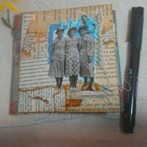 May include: Mixed media art piece featuring a collage with vintage photographs of three women. The artwork incorporates text from old book pages, blue stitching, and gold accents. A black pen is placed next to the artwork.